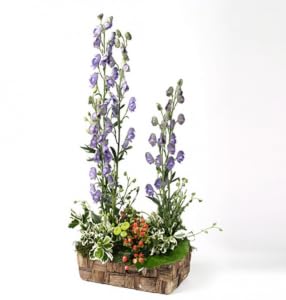 Arranjo floral vertical com flores lilases, flores pequenas e folhagem verde numa base retangular.